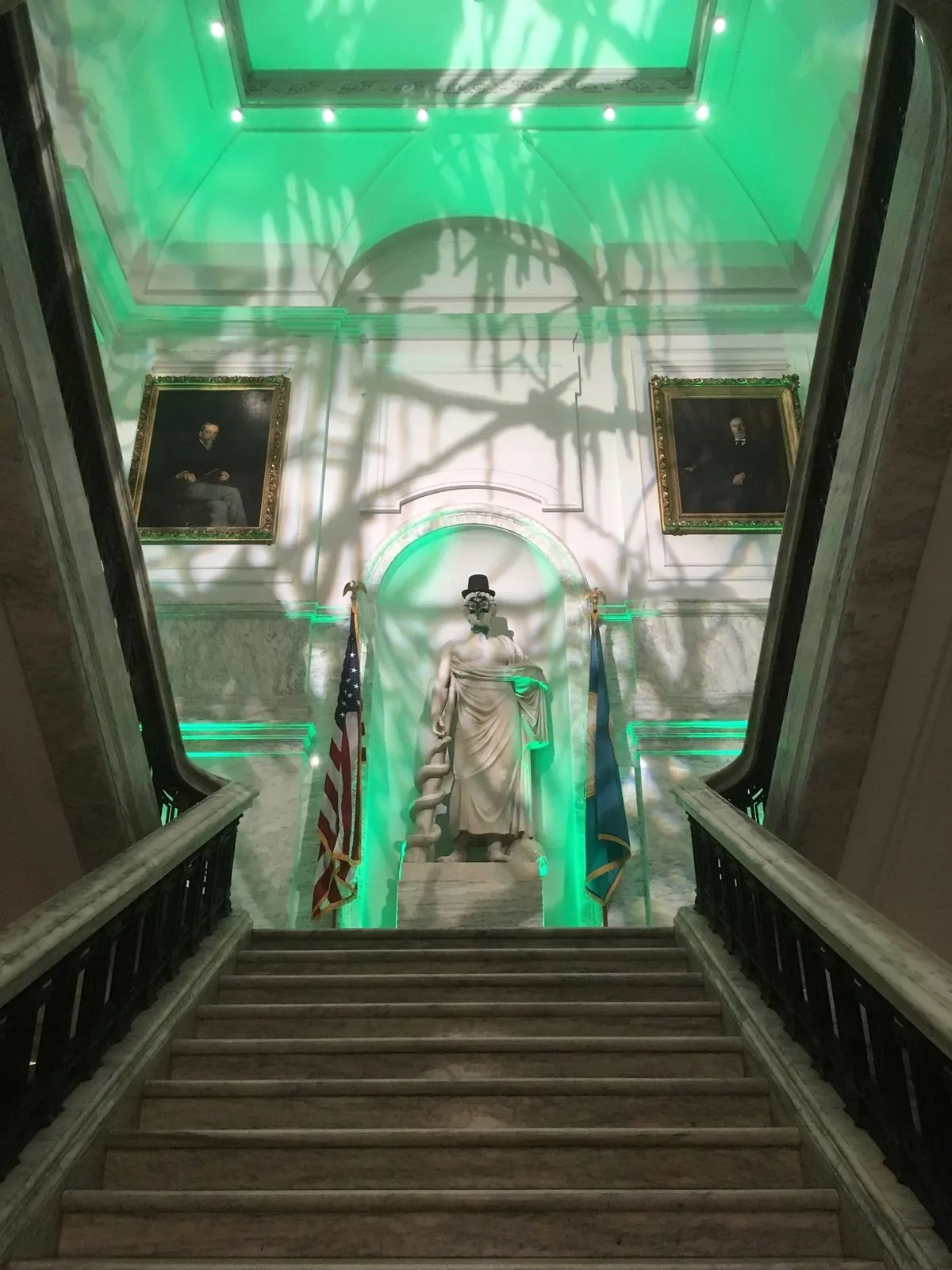 A marble staircase and statue inside a civic building washed in deep green uplighting.