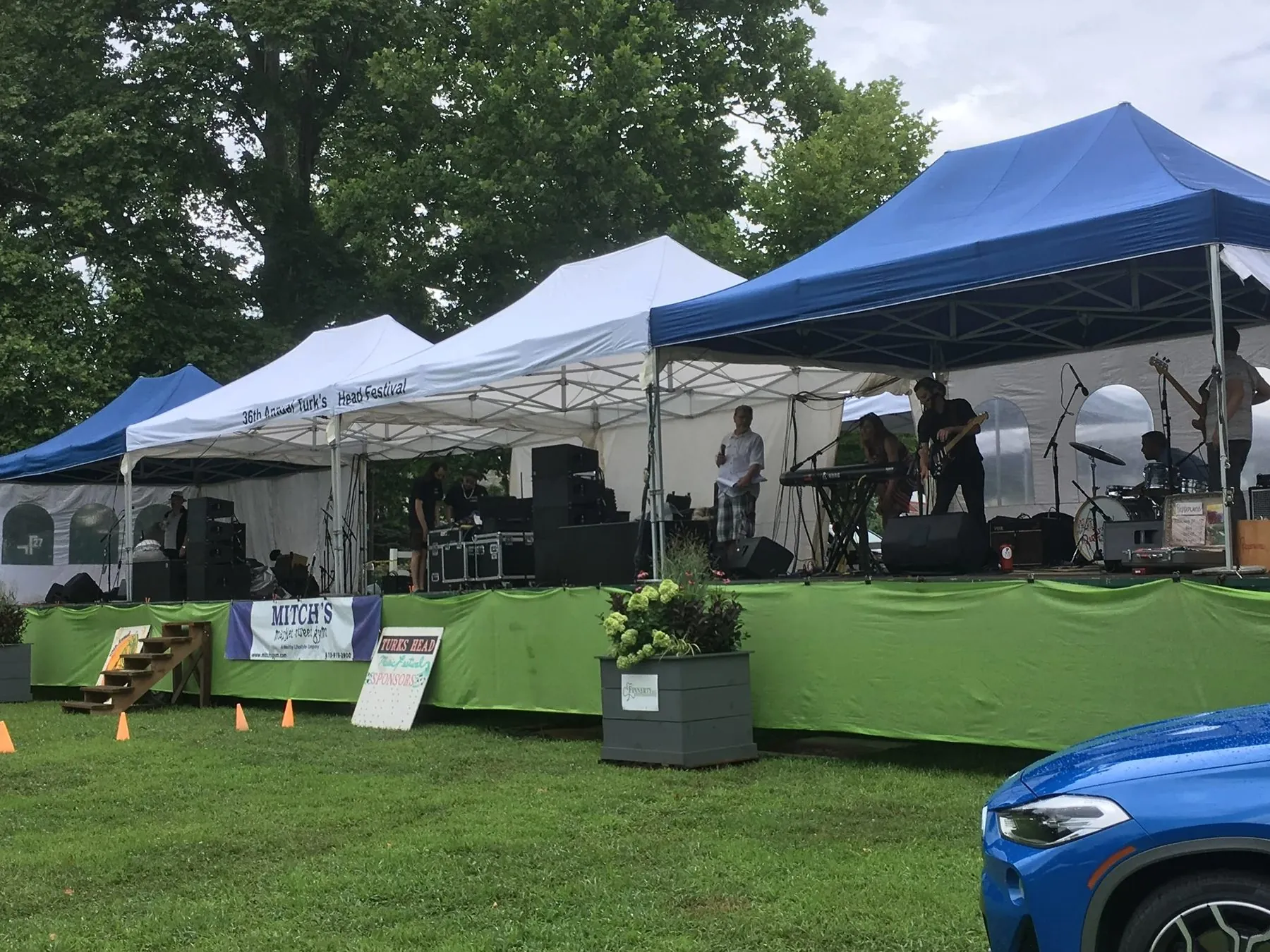 An outdoor festival stage under white and blue canopy tents with a live band performing.