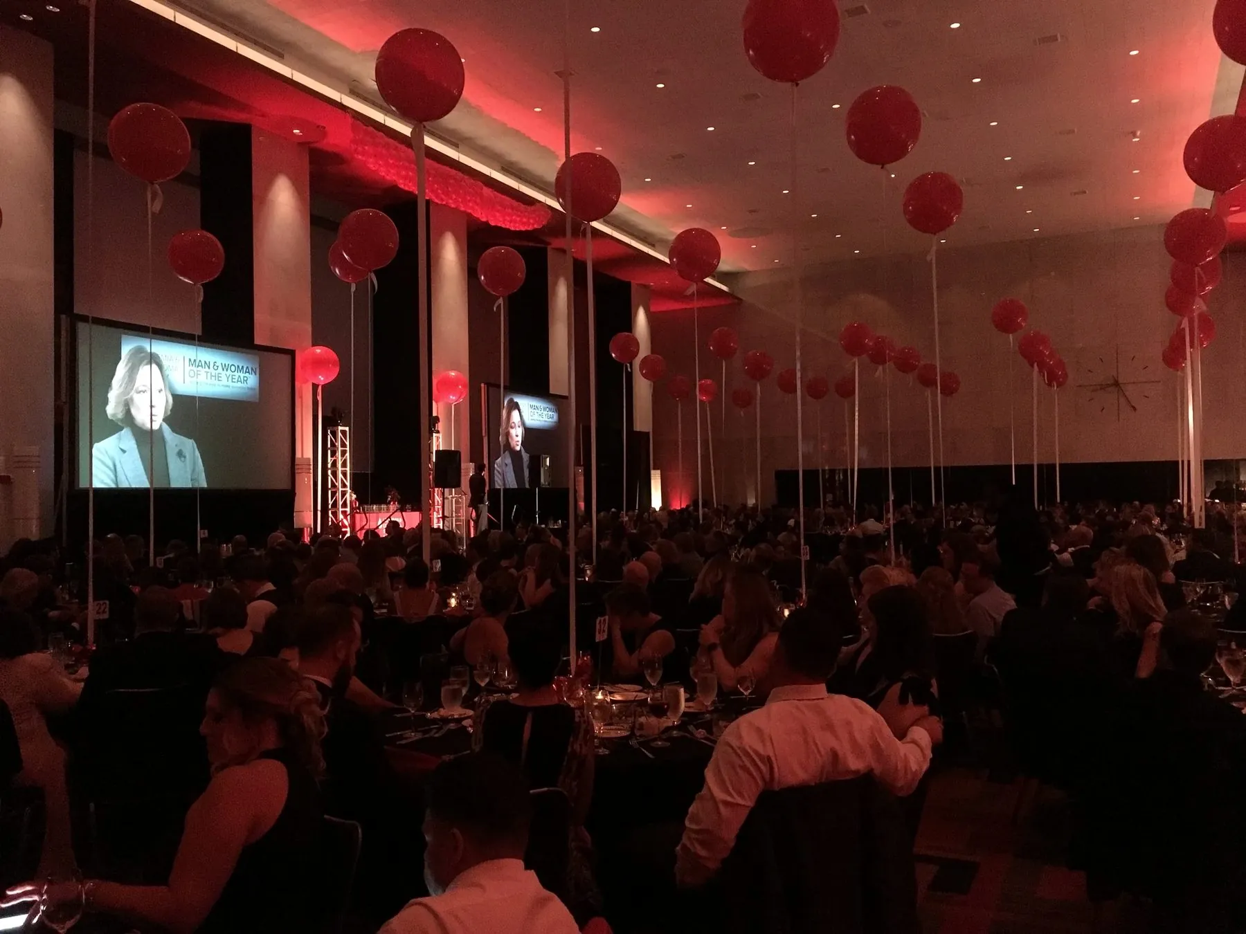 A packed gala ballroom lit in deep red, with red balloon clusters over the tables and the speaker projected on large screens.
