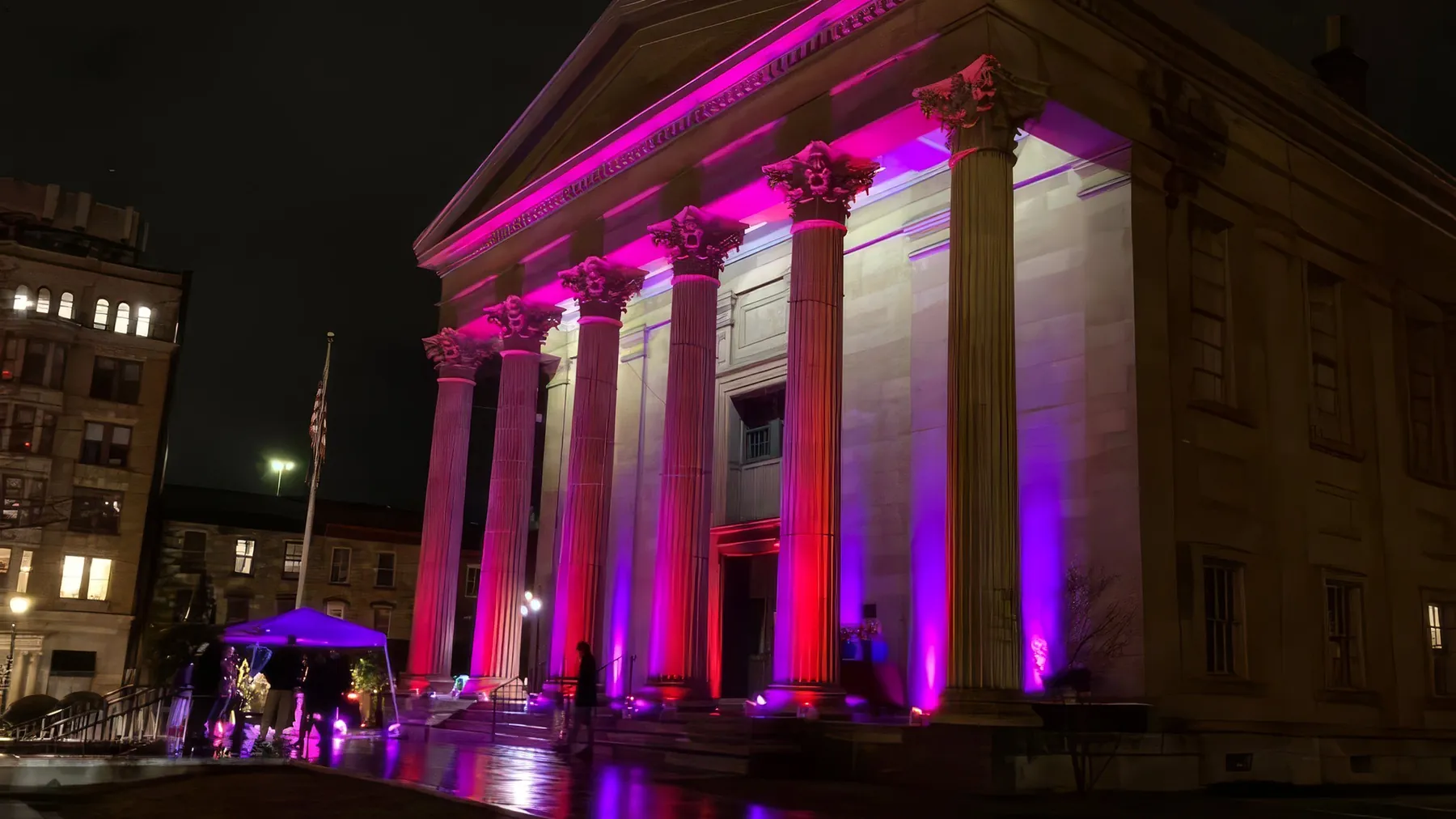 A neoclassical building lit at night in deep magenta and purple architectural uplighting, its columns and pediment washed in colour and reflected on the wet pavement below.