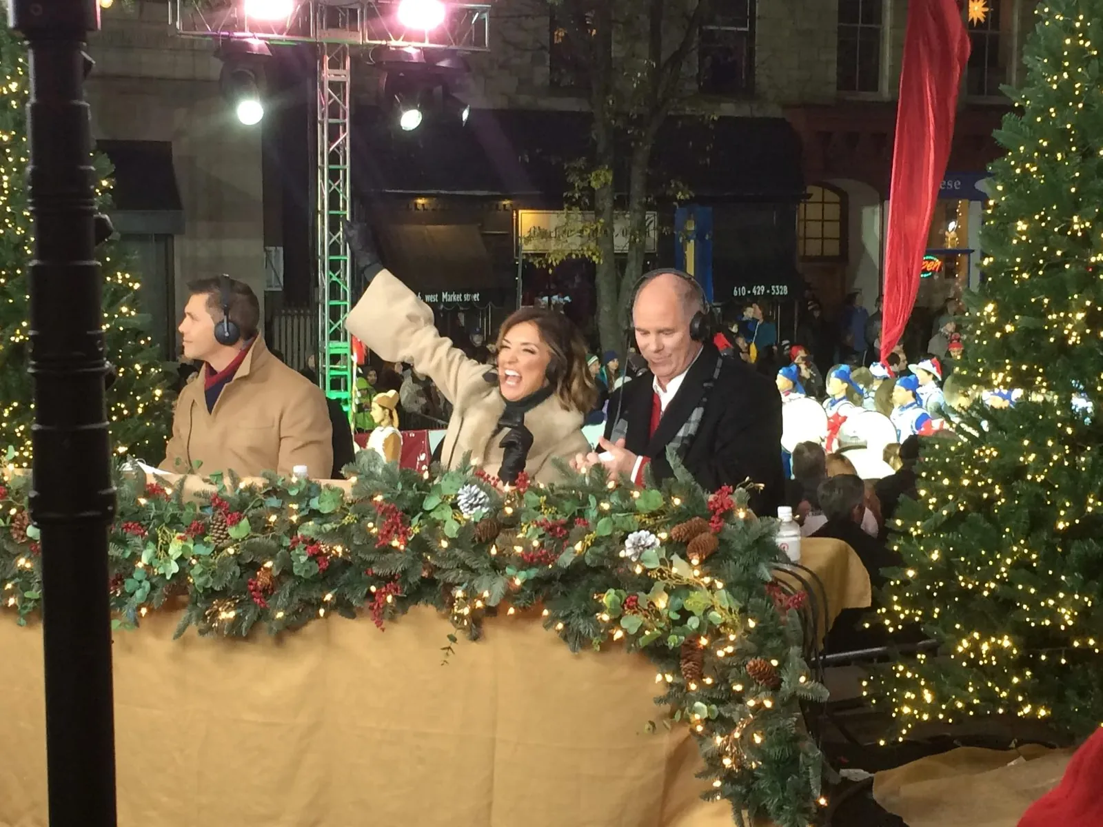A live broadcast position at an outdoor holiday event, with presenters in headsets at a garland-covered desk.