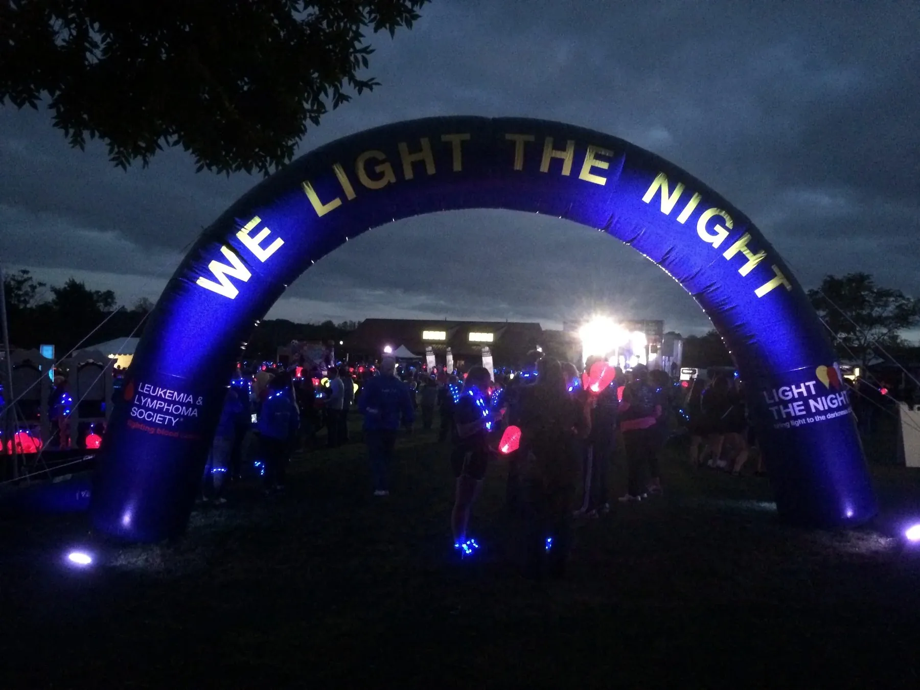 A large illuminated archway reading We Light The Night at a night-time charity walk, with a crowd gathered beneath it.