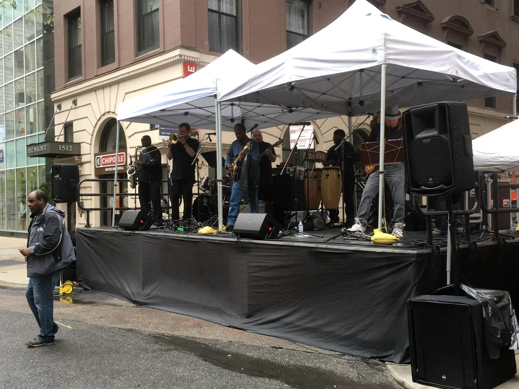 A live band performing on a covered street stage with monitors and loudspeakers in a Philadelphia city block.