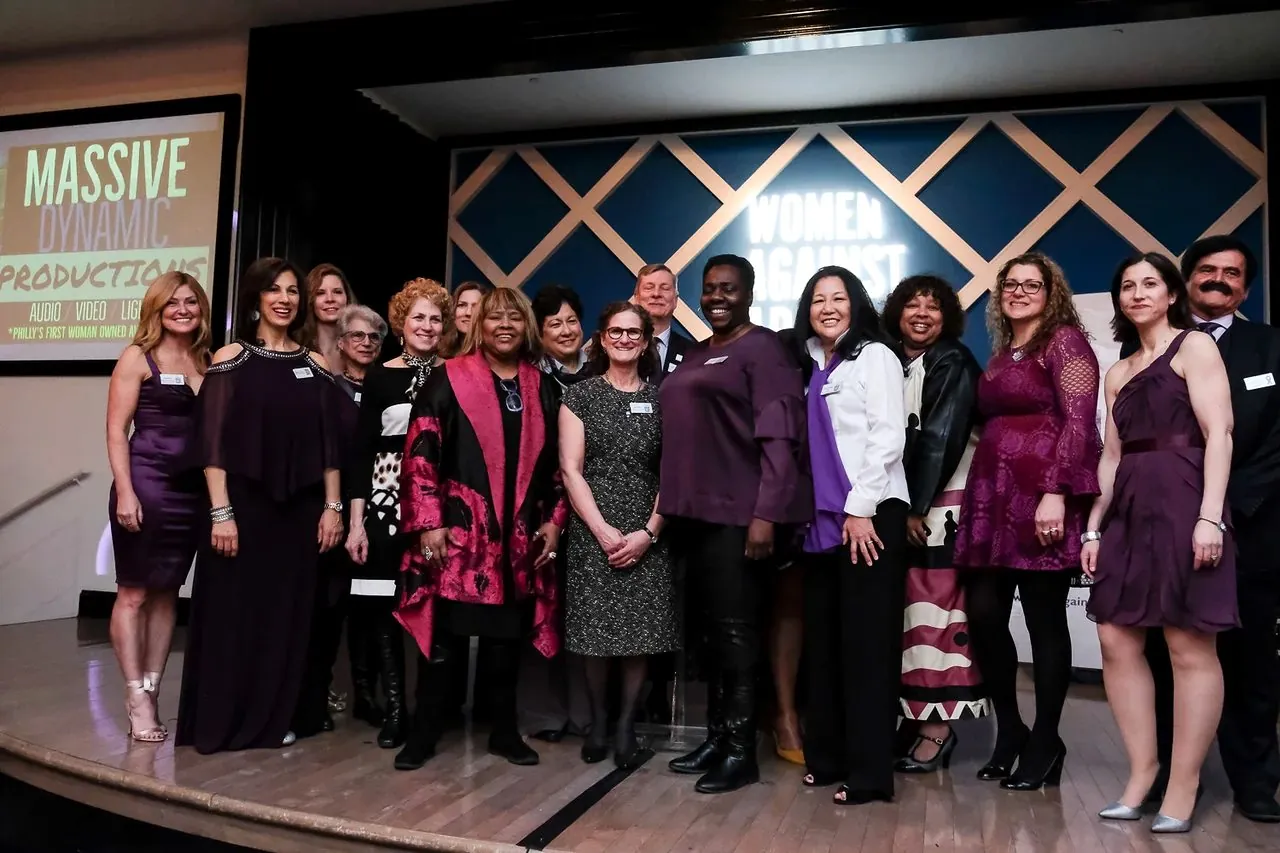 A group of honorees standing together on stage at the Women Against Abuse event, with the MDP logo on the screen behind them.